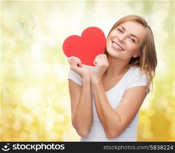 happiness, health, people, holidays and love concept - smiling young woman in white t-shirt holding red heart over yellow lights background