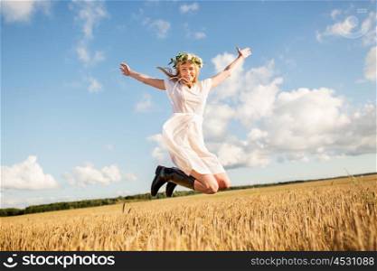 happiness, nature, summer holidays, vacation and people concept - smiling young woman in wreath of flowers and gumboots jumping on cereal field