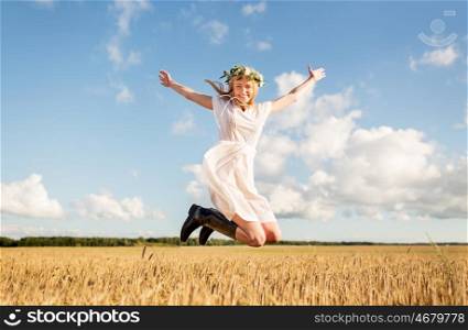 happiness, nature, summer holidays, vacation and people concept - smiling young woman in wreath of flowers and gumboots jumping on cereal field