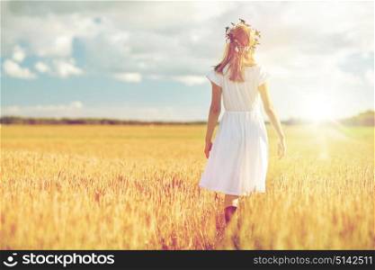 happiness, nature, summer holidays, vacation and people concept - smiling young woman in wreath of flowers and white dress walking along cereal field. happy young woman in flower wreath on cereal field
