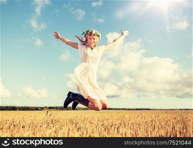 happiness, nature, summer holidays, vacation and people concept - smiling young woman in wreath of flowers and gumboots jumping on cereal field. happy woman in wreath jumping on cereal field