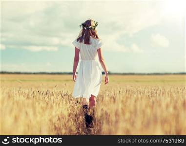 happiness, nature, summer holidays, vacation and people concept - smiling young woman in wreath of flowers and white dress walking along cereal field. happy young woman in flower wreath on cereal field