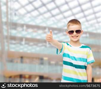 happiness, summer, childhood, gesture and people concept - smiling boy in sunglasses over shopping center background showing thumbs up