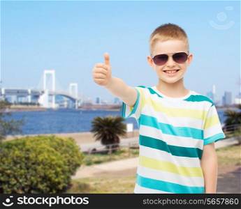 happiness, summer, childhood, gesture and people concept - smiling cute little boy in sunglasses showing thumbs up over city background