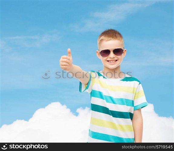 happiness, summer, childhood, gesture and people concept - smiling cute little boy in sunglasses over blue sky background showing thumbs up