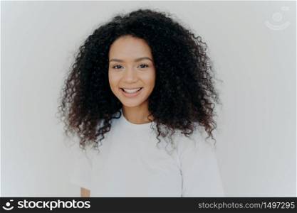 Happiness, wellbeing and positive emotions concept. Headshot of pleasant looking lady with crisp hair, smiles and laughs, has healthy skin, wears casual t shirt, models against white studio wall.