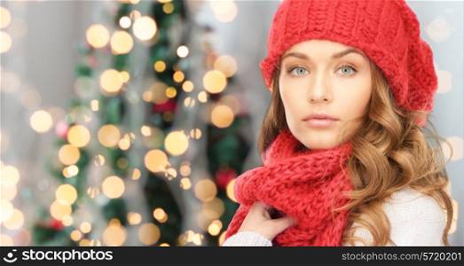 happiness, winter holidays and people concept - close up of young woman in red hat and scarf over christmas tree lights background