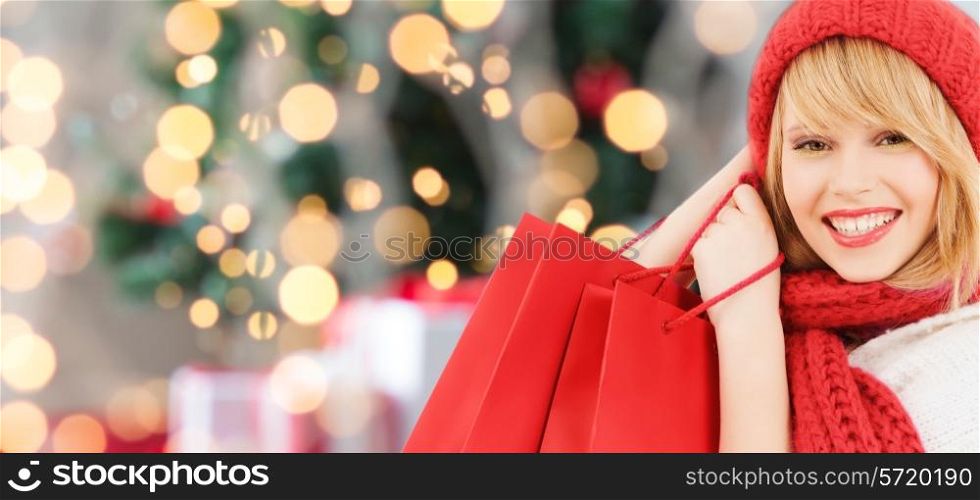 happiness, winter holidays and people concept - smiling young woman in hat and scarf with red shopping bags over christmas tree background
