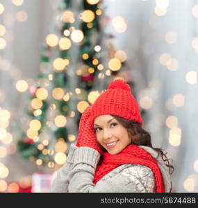 happiness, winter holidays and people concept - smiling young woman in red hat, scarf and mittens over christmas tree lights background