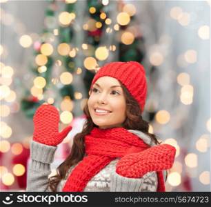 happiness, winter holidays and people concept - smiling young woman in red hat, scarf and mittens over christmas tree lights background