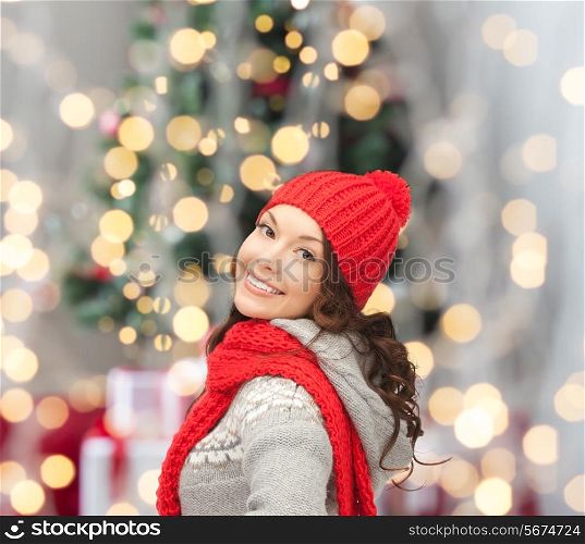 happiness, winter holidays and people concept - smiling young woman in red hat and scarf over christmas tree lights background
