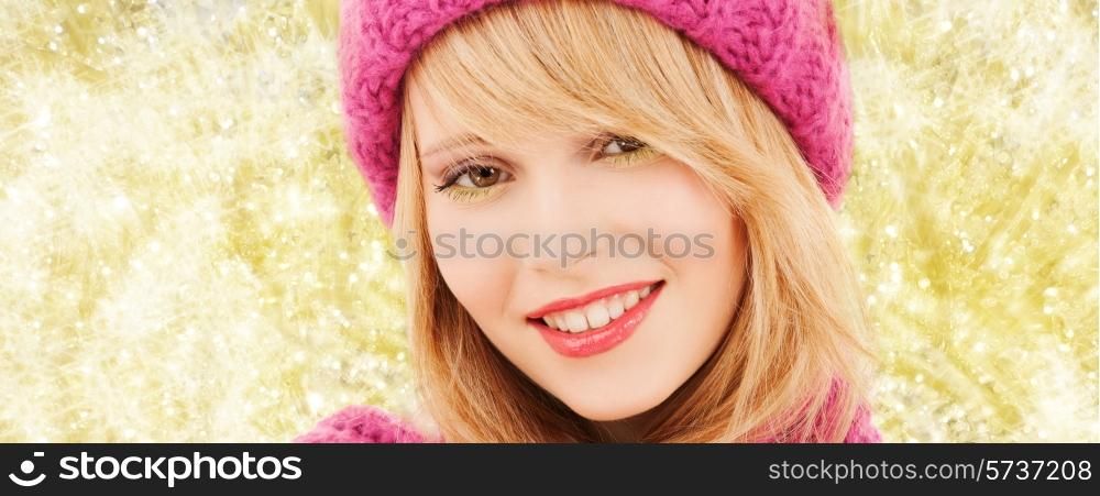 happiness, winter holidays, christmas and people concept - close up of smiling young woman in pink hat and scarf over yellow lights background