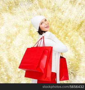 happiness, winter holidays, christmas and people concept - smiling young woman in white hat and mittens with red shopping bags over yellow lights background
