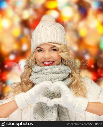 happiness, winter holidays, christmas and people concept - smiling young woman in white hat and mittens showing heart-shape gesture over shiny lights background