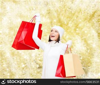 happiness, winter holidays, christmas and people concept - smiling young woman in white hat and mittens with red shopping bags over yellow lights background
