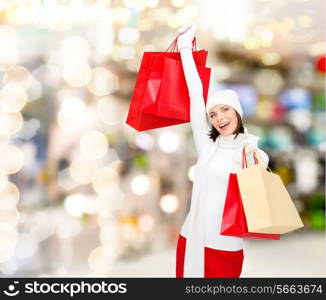 happiness, winter holidays, christmas and people concept - smiling young woman in white hat and mittens with red shopping bags over lights background