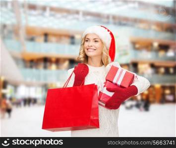happiness, winter holidays, christmas and people concept - smiling young woman in santa helper hat with gift boxes and shopping bag over shopping center background