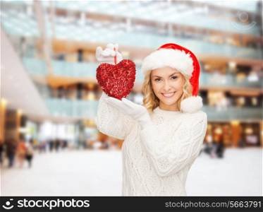 happiness, winter holidays, christmas and people concept - smiling young woman in santa helper hat with red heart decoration over shopping center background
