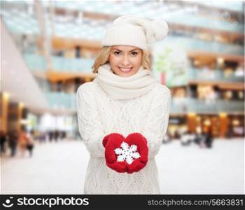 happiness, winter holidays, christmas and people concept - smiling young woman in hat, scarf and mittens holding snowflake decoration over shopping center background