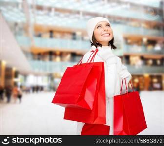 happiness, winter holidays, christmas and people concept - smiling young woman in white hat and mittens with red shopping bags over shopping center background