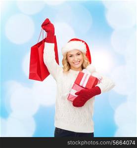 happiness, winter holidays, christmas and people concept - smiling young woman in santa helper hat with gifts and shopping bag over blue lights background