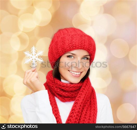 happiness, winter holidays, christmas and people concept - smiling young woman in red hat, scarf and mittens holding snowflake over beige lights background
