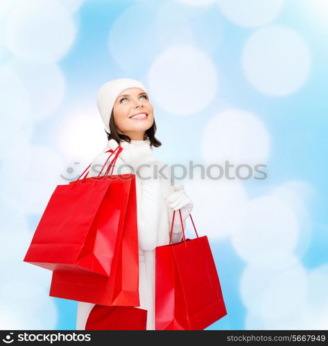 happiness, winter holidays, christmas and people concept - smiling young woman in white hat and mittens with red shopping bags over blue lights background