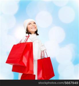 happiness, winter holidays, christmas and people concept - smiling young woman in white hat and mittens with red shopping bags over blue lights background