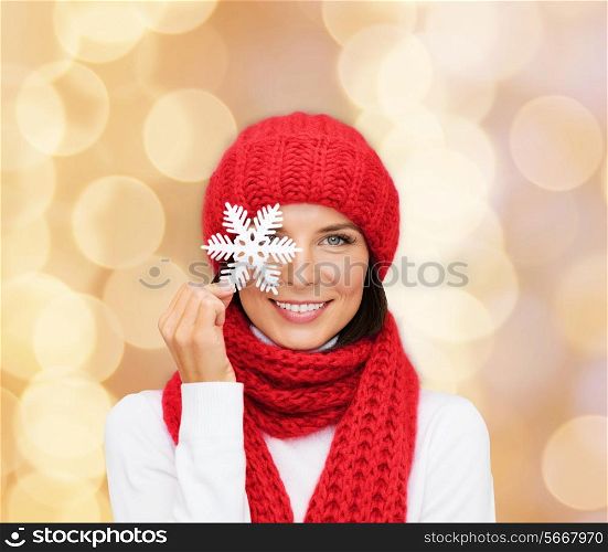 happiness, winter holidays, christmas and people concept - smiling young woman in red hat, scarf and mittens holding snowflake over beige lights background