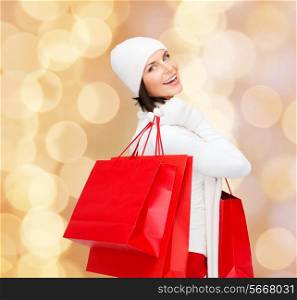 happiness, winter holidays, christmas and people concept - smiling young woman in white hat and mittens with red shopping bags over beige lights background