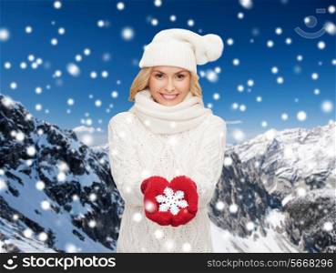 happiness, winter holidays, christmas and people concept - smiling young woman in hat, scarf and mittens holding snowflake decoration over snowy mountains background
