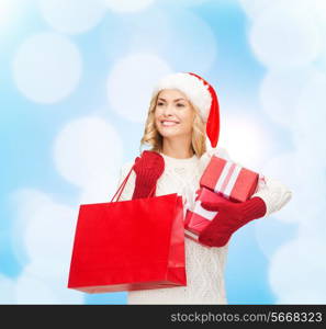 happiness, winter holidays, christmas and people concept - smiling young woman in santa helper hat with gift boxes and shopping bag over blue lights background