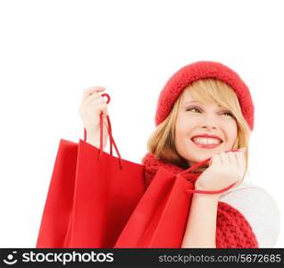 happiness, winter holidays, christmas and people concept - smiling young woman in hat and scarf with red shopping bags over white background