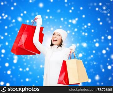 happiness, winter holidays, christmas and people concept - smiling young woman in white hat and mittens with red shopping bags over blue snowing background