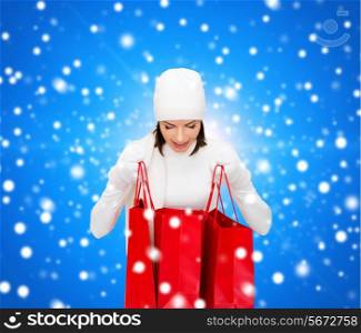 happiness, winter holidays, christmas and people concept - smiling young woman in white hat and mittens with red shopping bags over blue snowing background