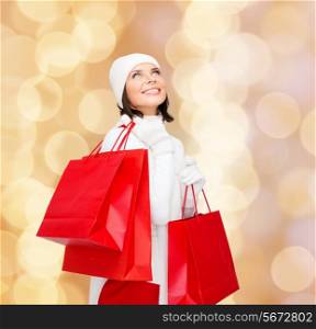 happiness, winter holidays, christmas and people concept - smiling young woman in white hat and mittens with red shopping bags over beige lights background