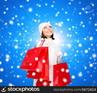 happiness, winter holidays, christmas and people concept - smiling young woman in white hat and mittens with red shopping bags over blue snowing background