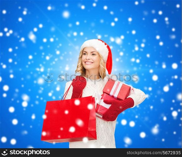 happiness, winter holidays, christmas and people concept - smiling young woman in santa helper hat with gift boxes and shopping bag over blue snowing background