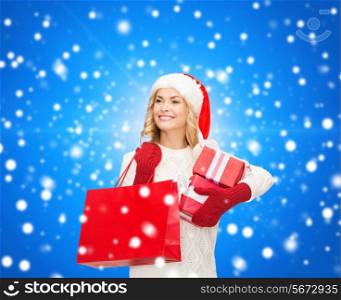 happiness, winter holidays, christmas and people concept - smiling young woman in santa helper hat with gift boxes and shopping bag over blue snowing background