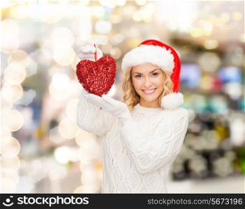 happiness, winter holidays, christmas and people concept - smiling young woman in santa helper hat with red heart decoration over lights background
