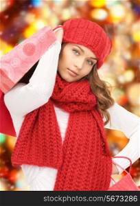 happiness, winter holidays, christmas and people concept - smiling young woman in hat and scarf with shopping bags over red lights background