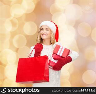 happiness, winter holidays, christmas and people concept - smiling young woman in santa helper hat with gift boxes and shopping bag over beige lights background