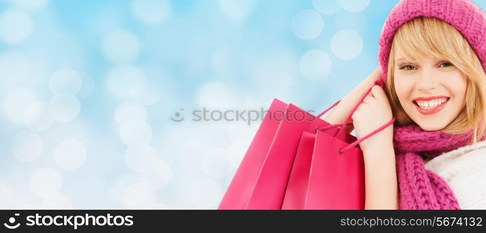 happiness, winter holidays, christmas and people concept - smiling young woman in hat and scarf with pink shopping bags over blue lights background