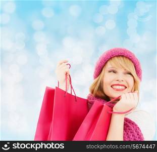 happiness, winter holidays, christmas and people concept - smiling young woman in hat and scarf with pink shopping bags over blue lights background