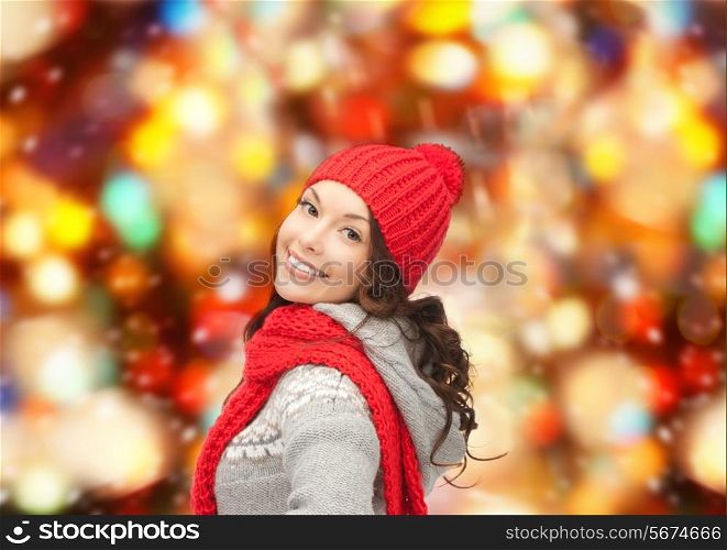 happiness, winter holidays, christmas and people concept - smiling young woman in red hat and scarf over red lights background