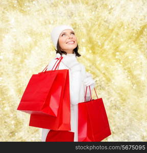 happiness, winter holidays, christmas and people concept - smiling young woman in white hat and mittens with red shopping bags over yellow lights background