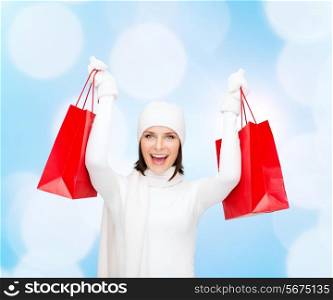 happiness, winter holidays, christmas and people concept - smiling young woman in white hat and mittens with red shopping bags over lights background