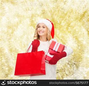 happiness, winter holidays, christmas and people concept - smiling young woman in santa helper hat with gift boxes and shopping bag over yellow lights background