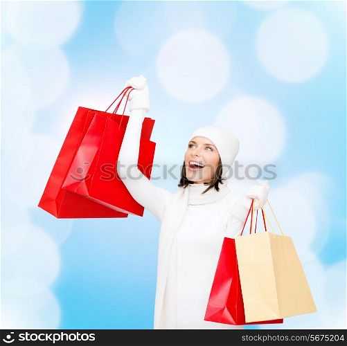 happiness, winter holidays, christmas and people concept - smiling young woman in white hat and mittens with red shopping bags over blue lights background
