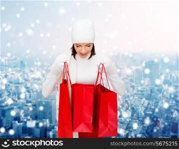 happiness, winter holidays, christmas and people concept - smiling young woman in white hat and mittens with red shopping bags over snowy city background